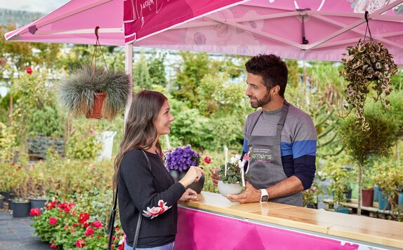 Verkaufsgespräch zwischen dem Verkäufer der Gärtnerei und einer Kundin unter einem pinken Faltpavillon mit Vordach.