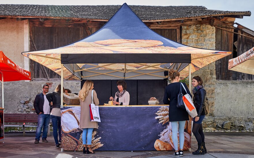 Das Marktzelt ist für eine Bäckerei und ist daher mit verschiedenem Brot bedruckt. Dahinter steht die Verkäuferin und schneidet ein Brot zur Verkostung an.