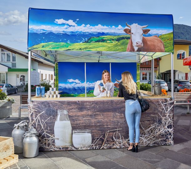 Der Faltpavillon ist für einen Milchhof bedruckt worden. Auf dem Marktzelt ist eine Kuh zu sehen und die Weide. Die Verkäuferin erklärt der Kundin die Milch.