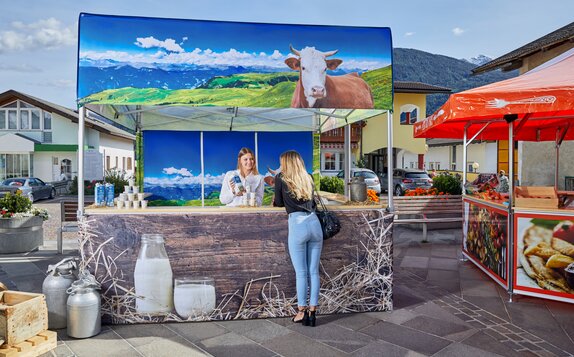 Der Faltpavillon ist für einen Milchhof bedruckt worden. Auf dem Marktzelt ist eine Kuh zu sehen und die Weide. Die Verkäuferin erklärt der Kundin die Milch.