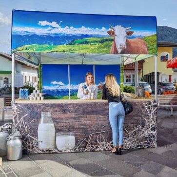 Der Faltpavillon ist für einen Milchhof bedruckt worden. Auf dem Marktzelt ist eine Kuh zu sehen und die Weide. Die Verkäuferin erklärt der Kundin die Milch.