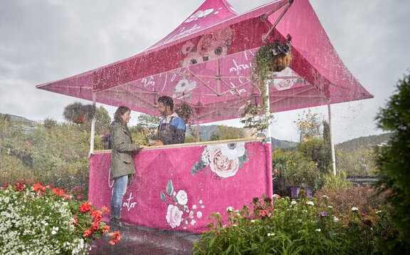 Bedruckter Faltpavillon mit Vordach im Regen. Die Kundin kauft beim Blumenhändler eine Blume. Es regnet und sie steht unter dem Vordach.