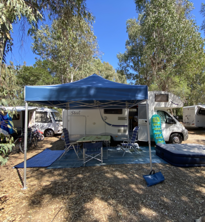 Blue camping tent 3x3 m stands in front of the caravan on the campsite. Under it there is a table with chairs.