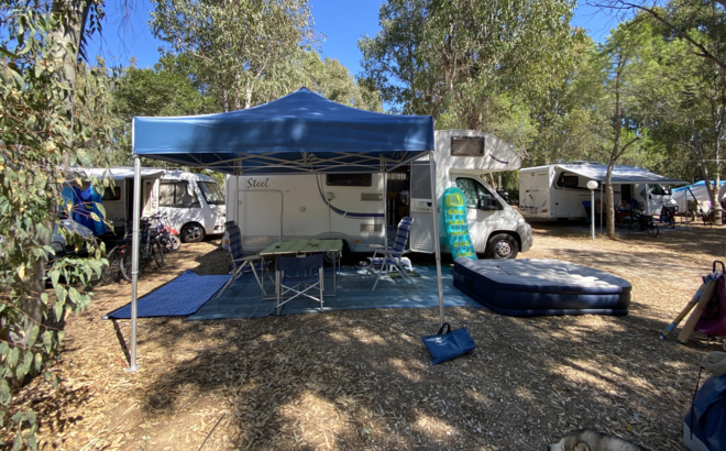 Gazebo da campeggio blu 3x3 m si trova di fronte alla roulotte del campeggio. Sotto di esso c'è un tavolo con sedie.