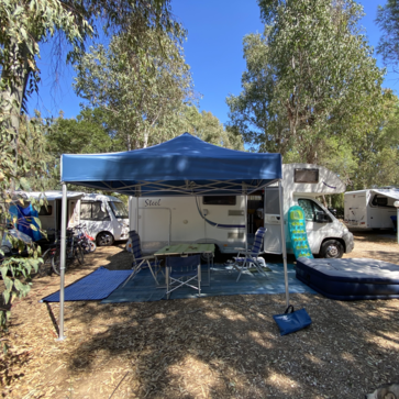 Blue camping tent 3x3 m stands in front of the caravan on the campsite. Under it there is a table with chairs.