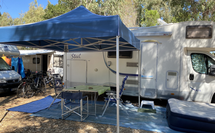 Blue camping tent 3x3 m stands in front of the caravan on the campsite. Under it there is a table with chairs.