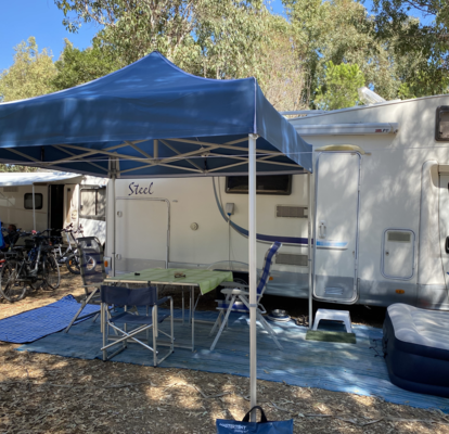 Blue camping tent 3x3 m stands in front of the caravan on the campsite. Under it there is a table with chairs.