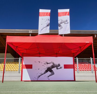 Roter Faltpavillon mit einer bedruckten Seitenwand und roter Struktur. Der Faltpavillon steht im Stadion, dahinter sieht man die Ränge.