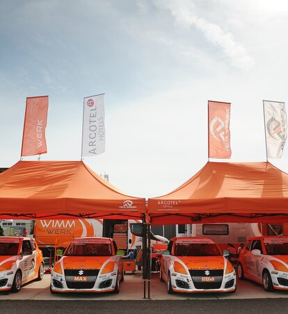 Zwei orange Paddockzelte vom Team Wimmer Werk stehen nebeneinander. Darunter stehen die Autos des Rennstalls.