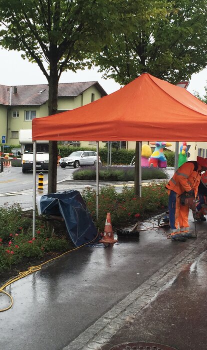 Un gazebo pieghevole arancione su un cantiere. Sotto il gazebo pieghevole ci sono due operai che lavorano.