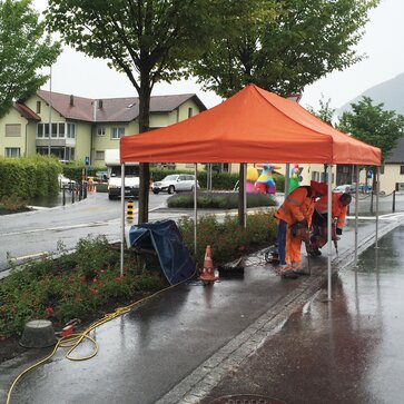 Ein oranger Faltpavillon steht auf einer Baustelle. Darunter befinden sich zwei Bauarbeiter, die Bohrarbeiten durchführen.