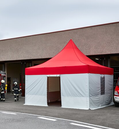 Feuerwehr Pavillon mit rotem Dach und grauen Seitenwänden steht vor dem Gebäude. Daneben stehen 2 Feuerwehrautos. Die Feuerwehrmänner kommen aus dem Gebäude.