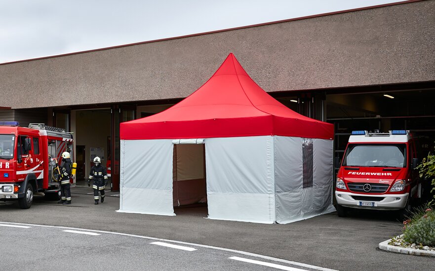 Feuerwehr Pavillon mit rotem Dach und grauen Seitenwänden steht vor dem Gebäude. Daneben stehen 2 Feuerwehrautos. Die Feuerwehrmänner kommen aus dem Gebäude.