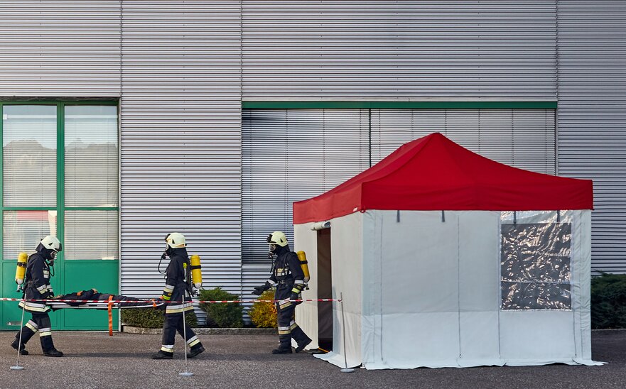 I vigili del fuoco portano la vittima dell´incidente nel gazebo pieghevole Rescue. Il gazebo Rescue ha un tetto rosso e pareti laterali grige.