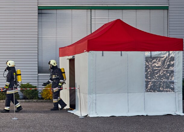 Die Feuerwehrmänner tragen das Unfallopfer in das Rescue-Zelt. Das Rescue-Zelt hat ein rotes Dach und graue Seitenwände.