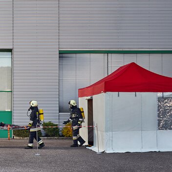 I vigili del fuoco portano la vittima dell´incidente nel gazebo pieghevole Rescue. Il gazebo Rescue ha un tetto rosso e pareti laterali grige.