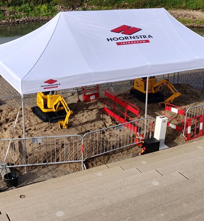 A white gazebo with the logo imprint "HOORNSTRA" is roofing the construction site. Underneath, two yellow excavators are working on the road maintenance. 