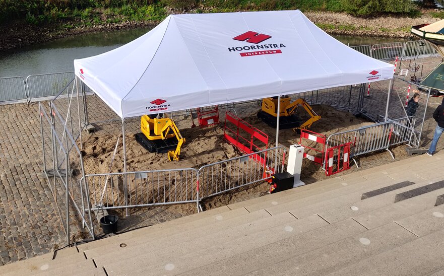 A white gazebo with the logo imprint "HOORNSTRA" is roofing the construction site. Underneath, two yellow excavators are working on the road maintenance. 