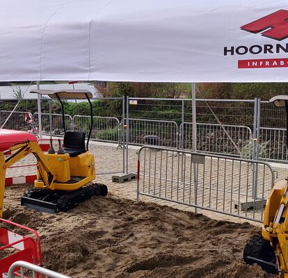 A white gazebo with the logo of "Hoornstra" is covering a construction site with yellow excavators standing on a sandy ground.