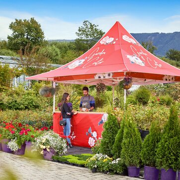 A gazebo with awnings is fully printed with flowers. It is located inbetween various plants of a garden market. Under the gazebo the seller is speaking with some clients. 