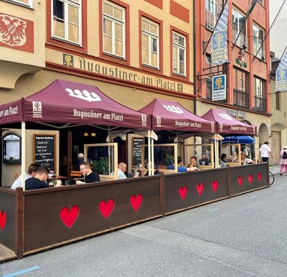 Gazebo pieghevole 3x1,5 bordeaux personalizzato con logo "Augustiner am Platzl". I gazebo sono nell'area esterna di un pub birreria ristorante e coprono i tavoli dove alcuni clienti stanno pranzando lungo le vie di una città