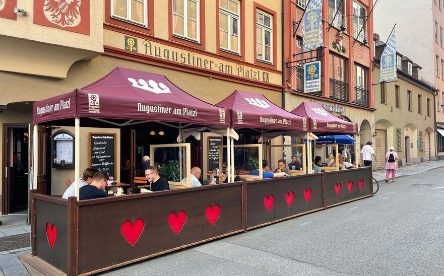 Gazebo pieghevole 3x1,5 bordeaux personalizzato con logo "Augustiner am Platzl". I gazebo sono nell'area esterna di un pub birreria ristorante e coprono i tavoli dove alcuni clienti stanno pranzando lungo le vie di una città