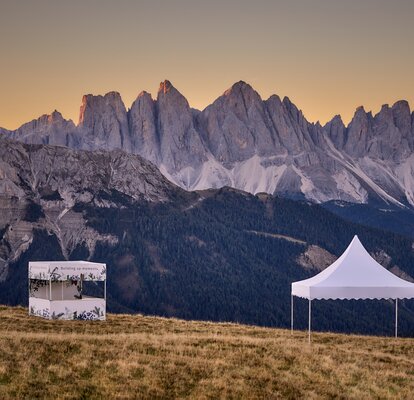 Ein bedrucktes weißes Faltzelt mit Flachdach und ein weißes Pagodenzelt stehen auf dem Plose-Berg. Die beiden Faltpavillons stehen im Sonnenuntergang vor einer Bergkette.