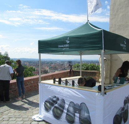 Gazebo pieghevole 2x2 m verde personalizzato con logo Svarovski Optik. Il gazebo ha tre pareti laterali stampate con delle foto di binocoli e con dei banconi per l'esposizione di prodotti e volantini. Il gazebo viene utilizzato per la promozione, ha una bandiera personalizzata e si trova in una terrazza con vista sulla città.