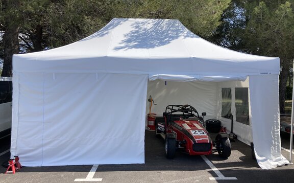 Auf dem Bild ist ein weißer, 6x4 m Faltpavillon mit Pavillon-Seitenteilen nach Maß zu sehen. Er besitzt eine Seitenwand mit 4 Türen, die individuell aufgerollt werden können. Darunter steht ein roter Rennwagen.