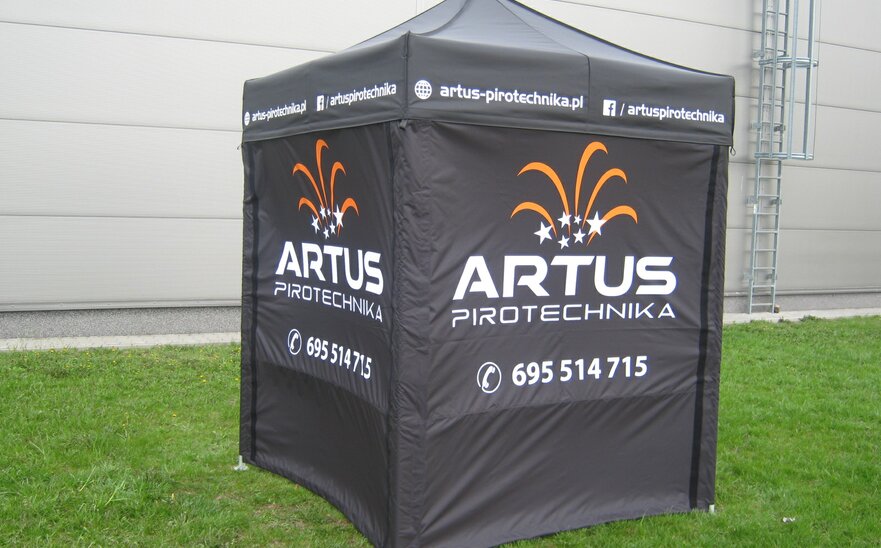 Una tenda promozionale nera di 2x2 m. Le pareti laterali sono stampate con il logo "Artus Pirotechnika" in bianco e in parte arancione, oltre che con il numero di telefono. La tenda si trova sul prato. 