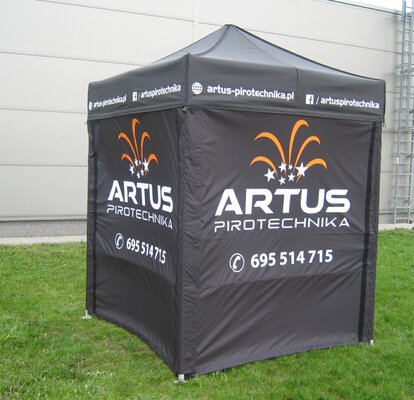 Una tenda promozionale nera di 2x2 m. Le pareti laterali sono stampate con il logo "Artus Pirotechnika" in bianco e in parte arancione, oltre che con il numero di telefono. La tenda si trova sul prato. 