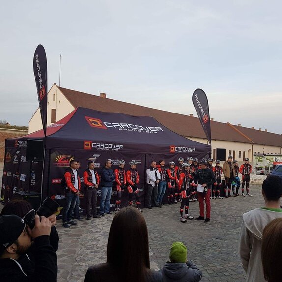 Una tenda di 8x4 m con pareti laterali chiuse e stampate di colore blu scuro. La tenda è stampata con il logo "Carcover Racing Team". Sul tetto a sinistra e a destra sono stampate le bandiere. Tutta la squadra è in piedi davanti alla tenda. 