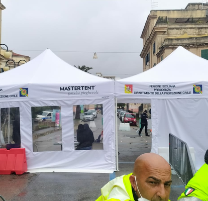 Due gazebo pieghevoli bianchi stanno uno accanto all'altro su una strada, hanno pareti laterali bianche con porte e finestre, sullo sfondo ci sono edifici e in primo piano due funzionari della protezione civile con gilet gialli.