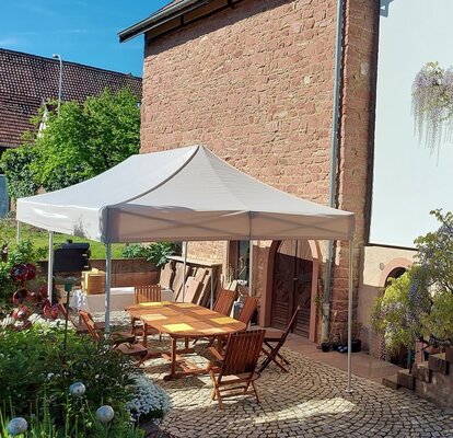 Das Gartenzelt 6x3 m in beige/ecru steht auf der Terrasse im Garten. Darunter steht eine Sitzecke mit einem großen Tisch und bequemen Stühlen. 