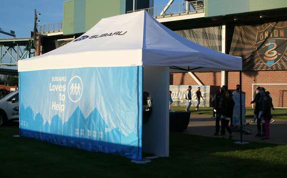 A Subaru folding gazebo with white roof and blue sidewall is standing on a lawn. In the background there are some people.