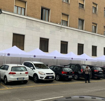 Ci sono nove gazebo con tetti bianchi davanti a un edificio. Davanti ai gazebo ci sono delle macchine e si vedono quattro persone.