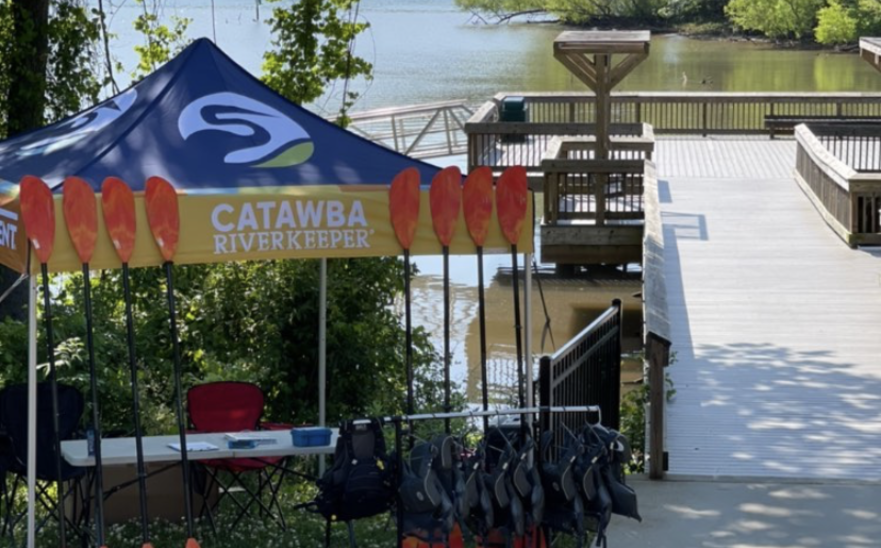 Ein Faltpavillon mit bedrucktem Dach steht am Ufer eines Flusses, umgeben von vielen Bäumen. Vier Kayaks liegen vor ihm in einer Wiese und an ihm angelehnt stehen acht Paddel. Unter dem Zelt sind ein kleiner Tisch und ein Stuhl.