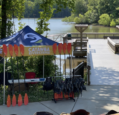 Ein Faltpavillon mit bedrucktem Dach steht am Ufer eines Flusses, umgeben von vielen Bäumen. Vier Kayaks liegen vor ihm in einer Wiese und an ihm angelehnt stehen acht Paddel. Unter dem Zelt sind ein kleiner Tisch und ein Stuhl.