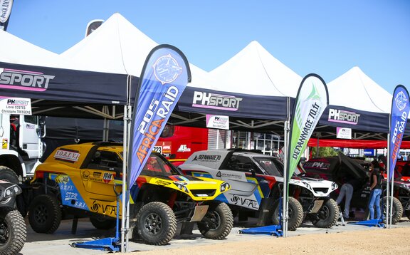 Four folding gazebos with white roof and blue valance are standing next to each other. There is a race car under each of them. In front of every tent there is a printed flag.