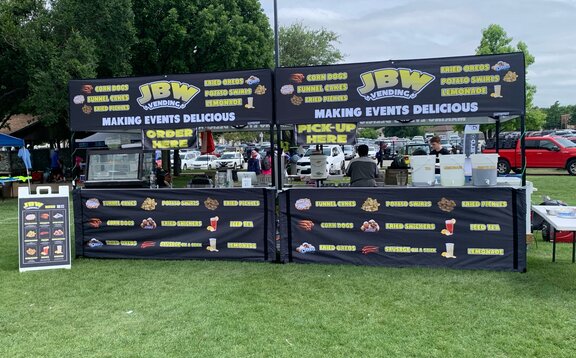 Two black, printed folding gazebos are standing next to each other on a lawn. They're stands where food and drinks are sold.