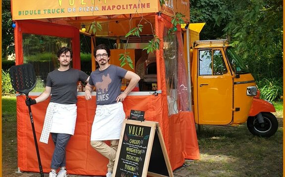 Ein oragner 2x2 Faltpavillon ist mit dem Firmennamen bedruckt. Der Pavillon hat Seitenwände. Vor dem Pavillon stehen zwei Personen mit Küchenbekleidung und Utensilien in der Hand. Der Pavillon steht vor dem mobilen Pizzaofen.