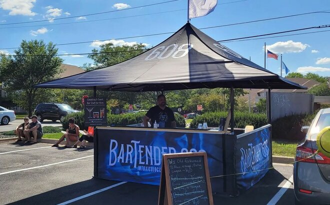 Sull'immagine c'è un gazebo nero con stampa, con un bancone in legno e una bandiera sul tetto. È un bar.