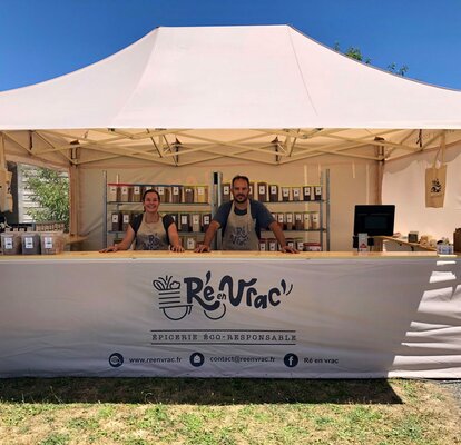 Sull'immagine c'è un gazebo pieghevole con tettoia, un bancone di legno e stampa. Sotto di esso ci sono due persone e vari prodotti. È un gazebo per mercati.