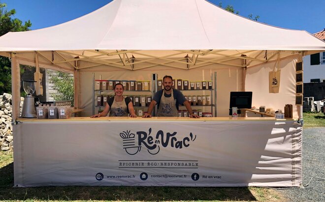 Sull'immagine c'è un gazebo pieghevole con tettoia, un bancone di legno e stampa. Sotto di esso ci sono due persone e vari prodotti. È un gazebo per mercati.