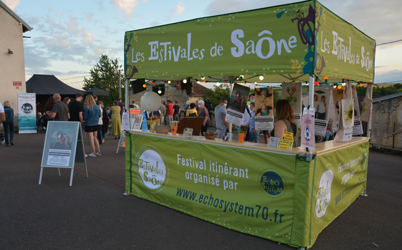 A green folding gazebo with a flat roof and a wooden counter can be seen. The tent is illuminated and people can be seen in the background.