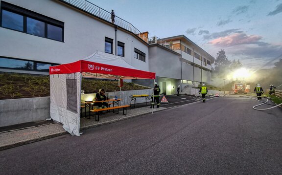 Neben einer Straße steht ein rot-weißes Feuerwehrzelt. Rund um das Zelt sind mehrere Feuerwehrmänner zu sehen und unter dem Zelt sitzt ein Feuerwehrmann auf einer Bank an einem Tisch.