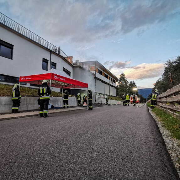 Next to a road there is a red and white fire brigade tent. Several firemen can be seen around the tent.