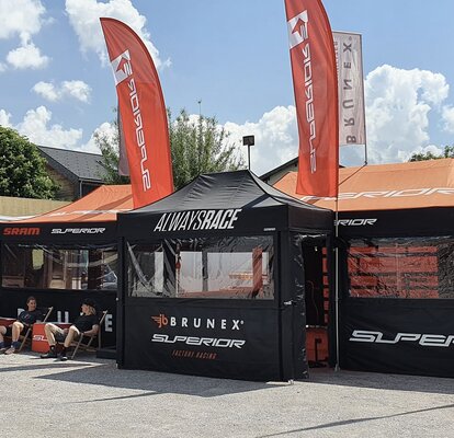 Es stehen drei Faltpavillons mit orangen Dächern und Flaggen auf einem Platz, links daneben ein Wohnwagen. Im Hintergrund befinden sich Bäume.