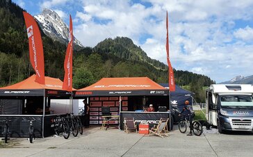 Es stehen zwei Faltpavillons mit orangen Dächern und Flaggen auf einem Platz, rechts daneben ein Wohnwagen. Im Hintergrund befinden sich Wälder und Berge.