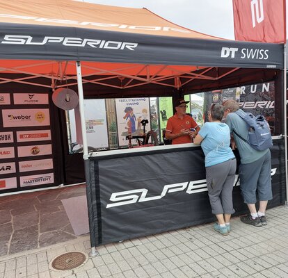 Auf dem Bild ist ein Faltpavillon mit Seitenwänden mit PVC Fenstern. An der Vorderseite hat es eine Seitenwand auf halber Höhe. Dort steht unter dem Zelt eine Person und vor dem Zelt zwei Personen. 